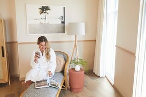 A person sitting on a patterned sofa in a room with a large window, a standing lamp, and a framed picture on the wall. - The Ryder Hotel (Charleston)