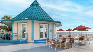 A small, single-story building with a blue roof and beige walls, featuring outdoor seating with red umbrellas and a view of the ocean.