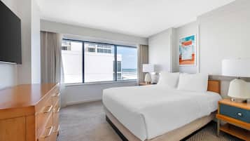A hotel room with a large bed, a wooden dresser, a flat-screen TV, a window with a view of buildings, and a painting on the wall.