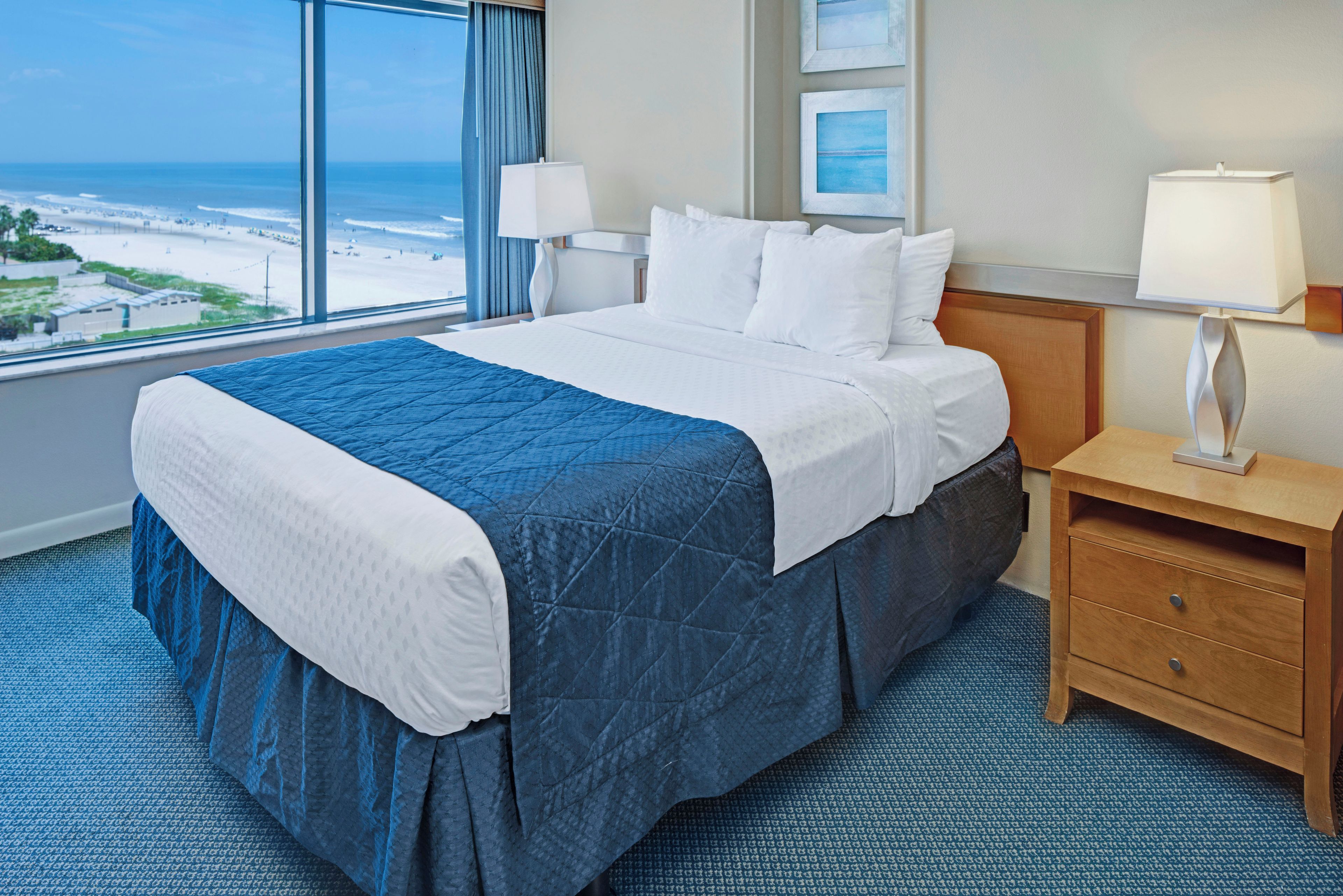 A hotel room with a large bed, a nightstand, a lamp, and a view of the beach.