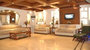 A spacious living room with a wooden ceiling, a large mirror, and a flat-screen TV mounted on the wall. - Hotel Emperor Palms (New Delhi)