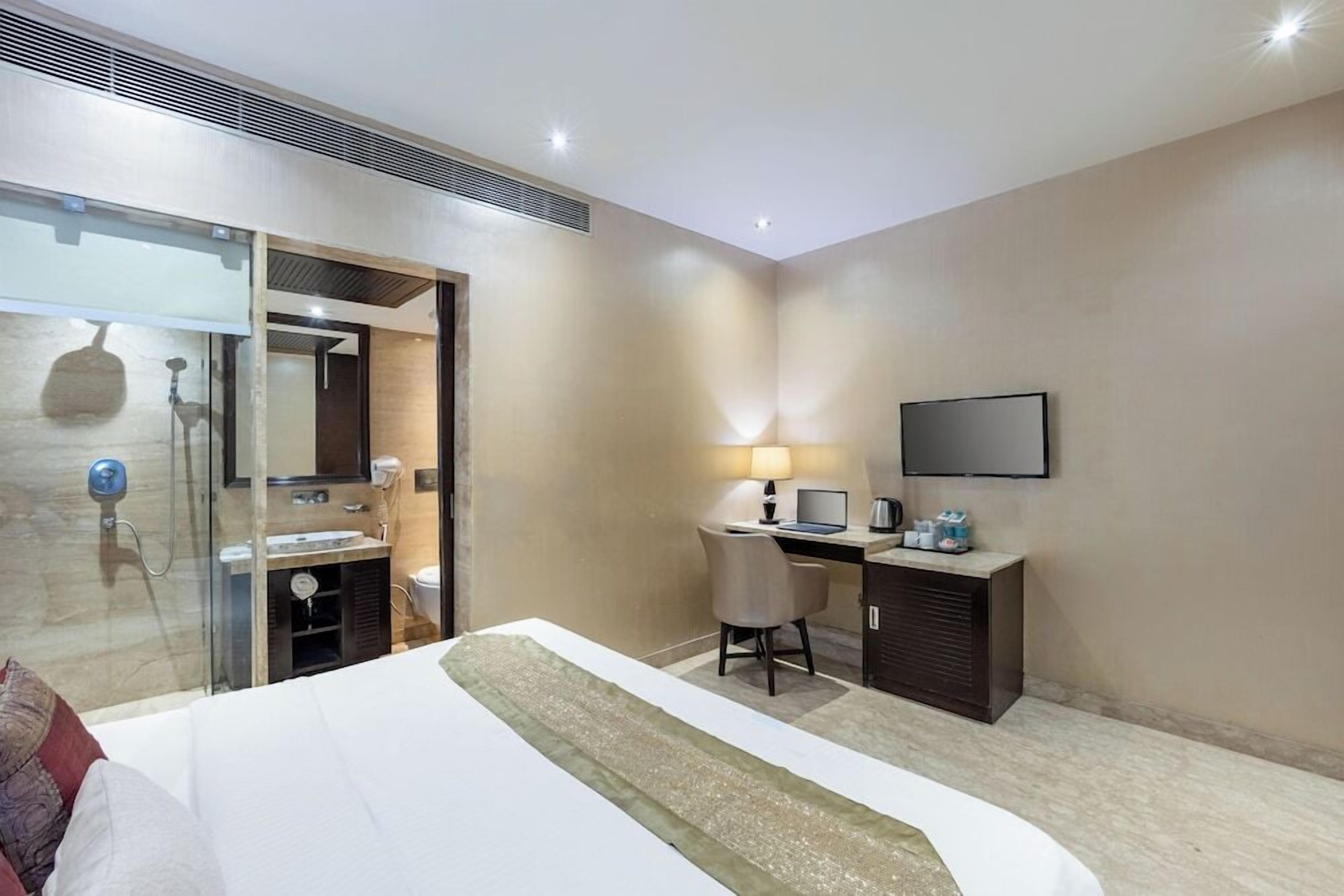 A hotel room with a bed, desk, chair, TV, and a glass shower area.