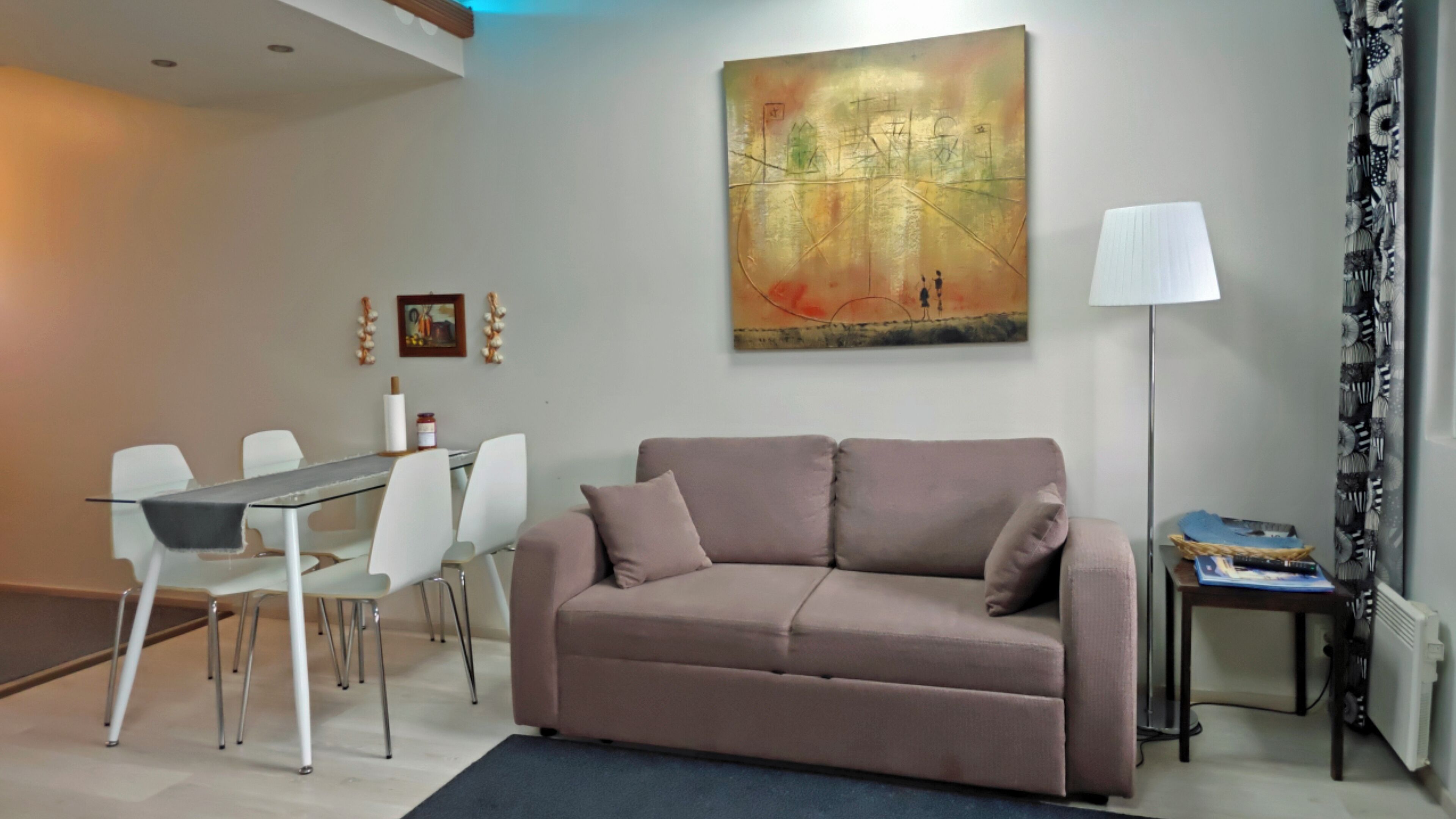 A living room with a sofa, a dining table with chairs, a painting on the wall, a lamp, and a radiator.