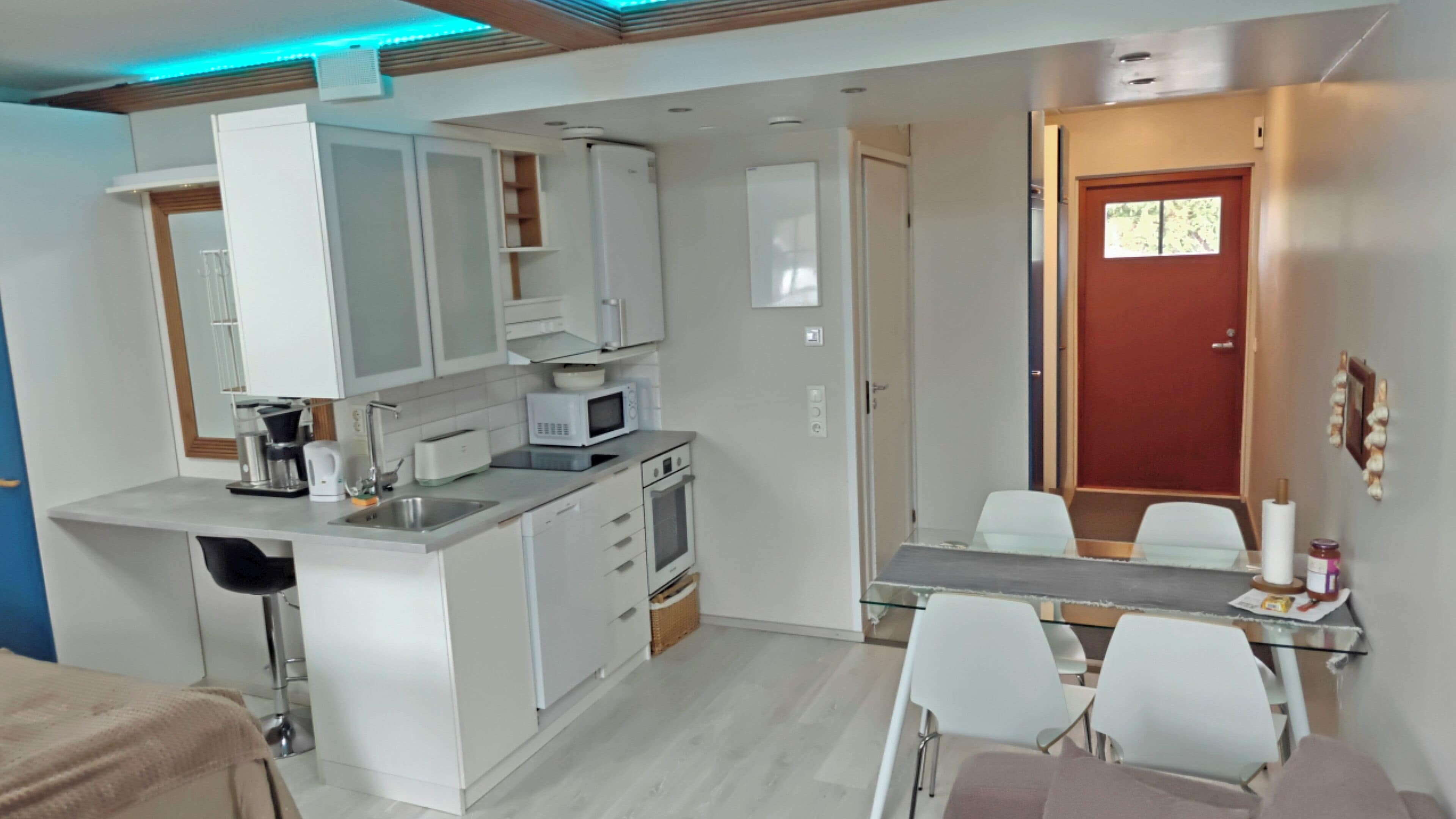 Una cocina compacta con armarios blancos, un microondas y un lavamanos. Hay una zona de comedor con una mesa y sillas.