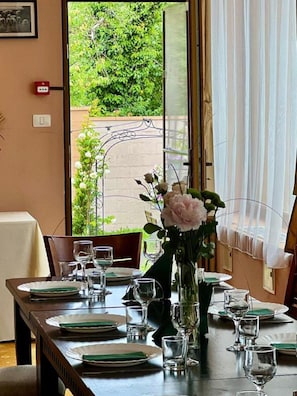 A dining table set for four with a vase of flowers, a window with a view of greenery, and a door with a red emergency light.