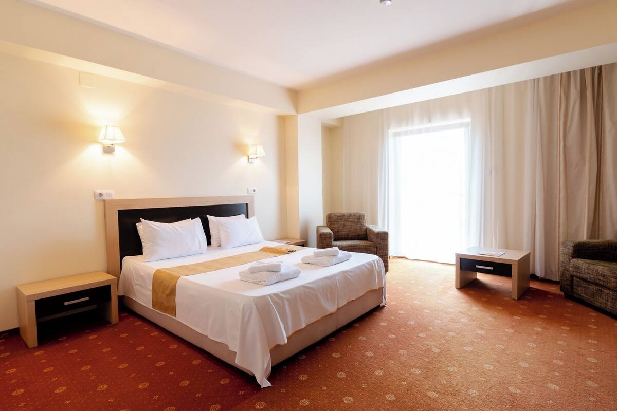 A hotel room with a bed, two bedside lamps, a chair, a small table, and a large window with curtains.