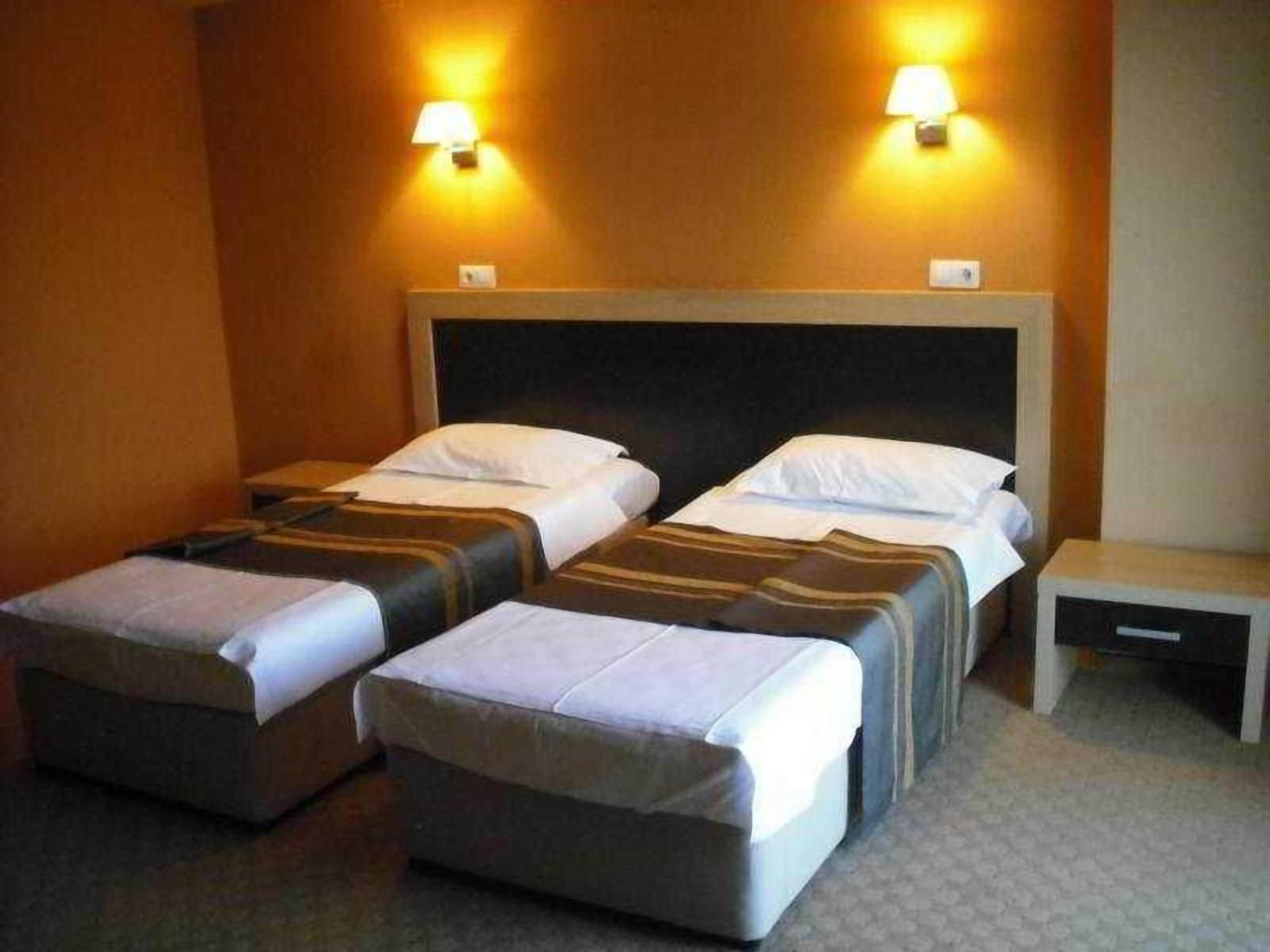 A hotel room with two beds, each with a pillow and a blanket, a bedside table, and two wall-mounted lamps.