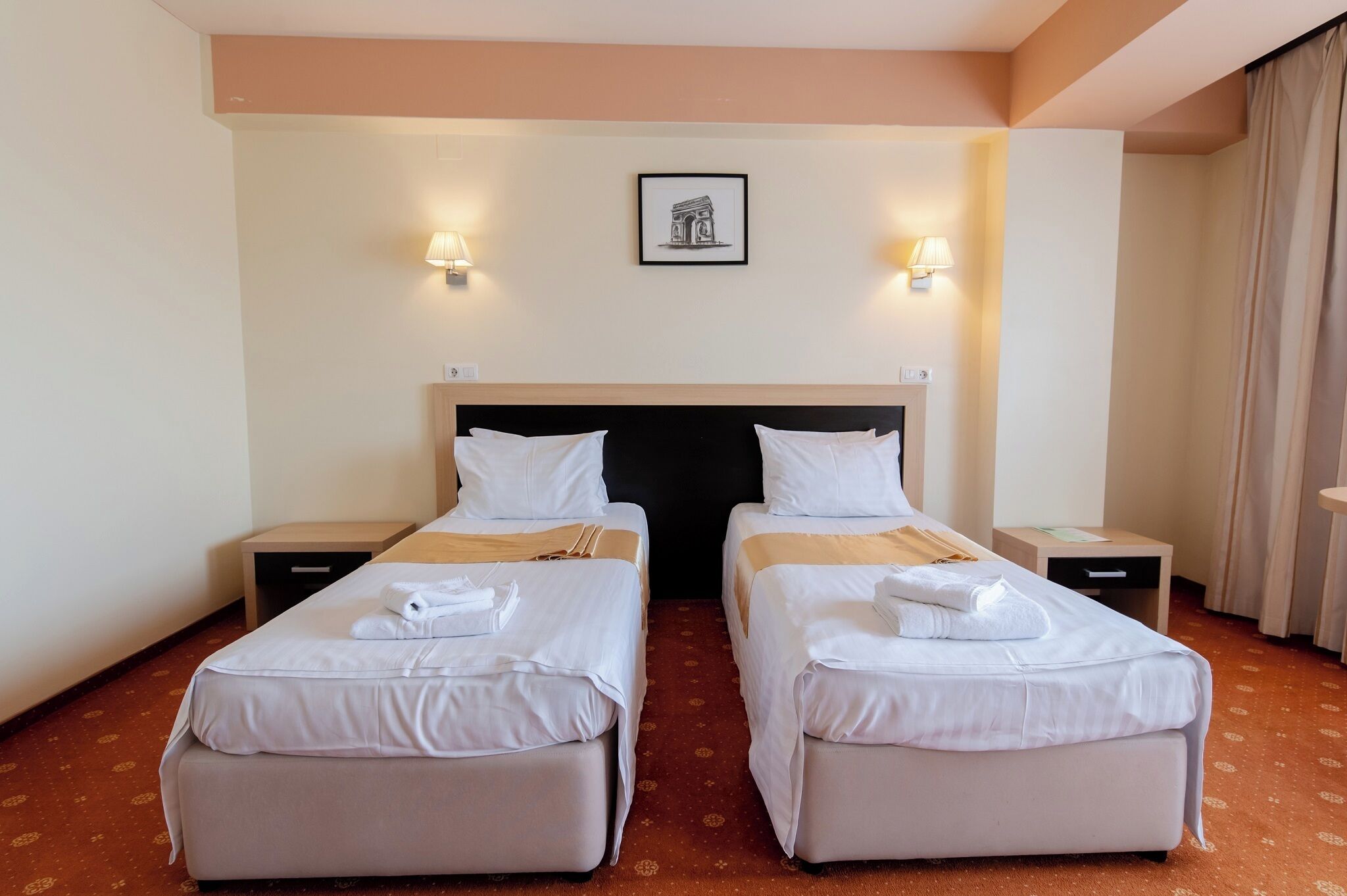 Habitación de hotel con dos camas individuales, un cabecero grande, mesitas de noche, lámparas fijadas a la pared y un cuadro enmarcado.