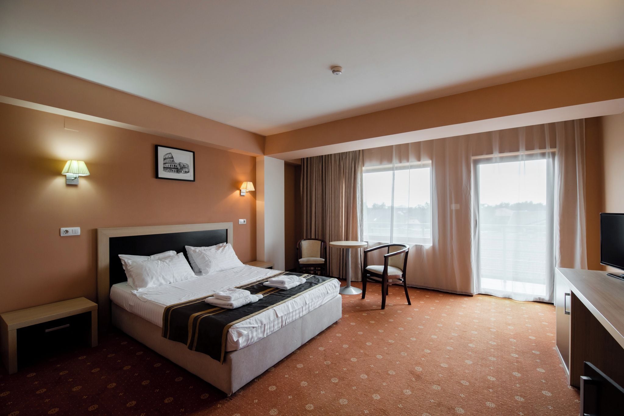 A hotel room with a bed, bedside lamps, a small table, a chair, a TV, and a window with curtains.