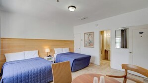 A hotel room with two beds, a wooden headboard, a small table with cups, and a bathroom visible through an open door.