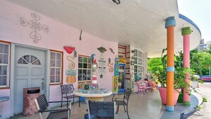 A colorful building with a pink facade, a covered outdoor seating area with chairs and a table, and large potted plants.