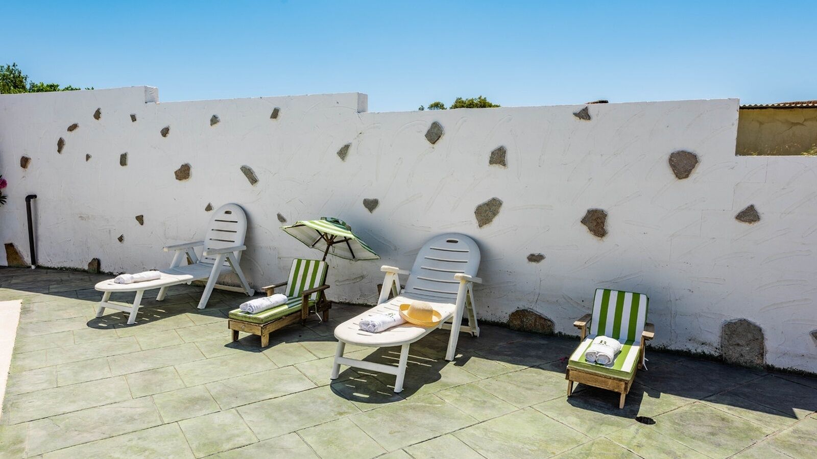 A patio area with five lounge chairs and a small table, each with a towel and a bottle, against a white wall with stone patches.