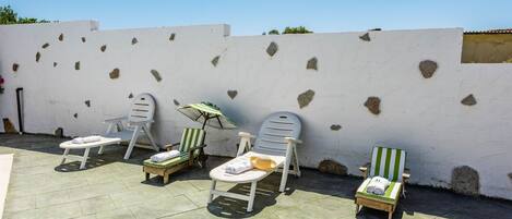 A patio area with five lounge chairs and a small table, each with a towel and a bottle, against a white wall with stone patches.