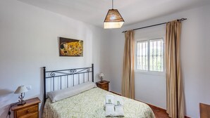 A bedroom with a bed, a nightstand, a lamp, a painting, and a window with curtains. - Chalet Algar by Ruralidays (Cádiz)