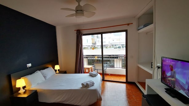 A hotel room with a bed, a TV, a ceiling fan, and a balcony with a view of buildings. - Hostal Balearic (Sant Antoni de Portmany)