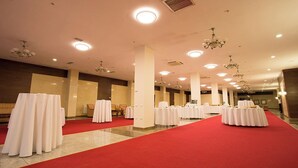 A spacious banquet hall with multiple round tables covered with white tablecloths, arranged on a red carpet.