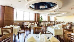 A spacious dining area with multiple tables and chairs, large windows, and a modern ceiling design.