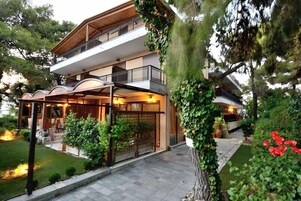 Un edificio de dos pisos con un balcón, una zona de estar cubierta al aire libre y un jardín bien cuidado con árboles y flores rojas.