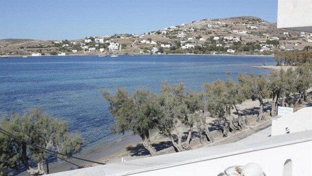 A coastal view with a clear blue sea, a sandy beach, and a white building with a balcony overlooking the water.