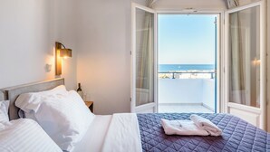 A hotel room with a bed, a nightstand, a bottle of wine, and a glass. There is a balcony with a view of the sea. - Marianna Hotel (Santorini)