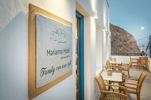 A Mariana Hotel sign with a building illustration, wooden furniture, and a view of a rocky coastline. - Marianna Hotel (Santorini)