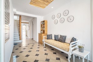 A modern interior with a checkered floor, a white sofa with patterned cushions, a wooden bookshelf, and a wall clock. - Marianna Hotel (Santorini)
