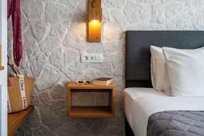 A bedroom with a stone wall, a bedside table with a book and sunglasses, a nightstand with a lamp, and a bed with white bedding. - Marianna Hotel (Santorini)