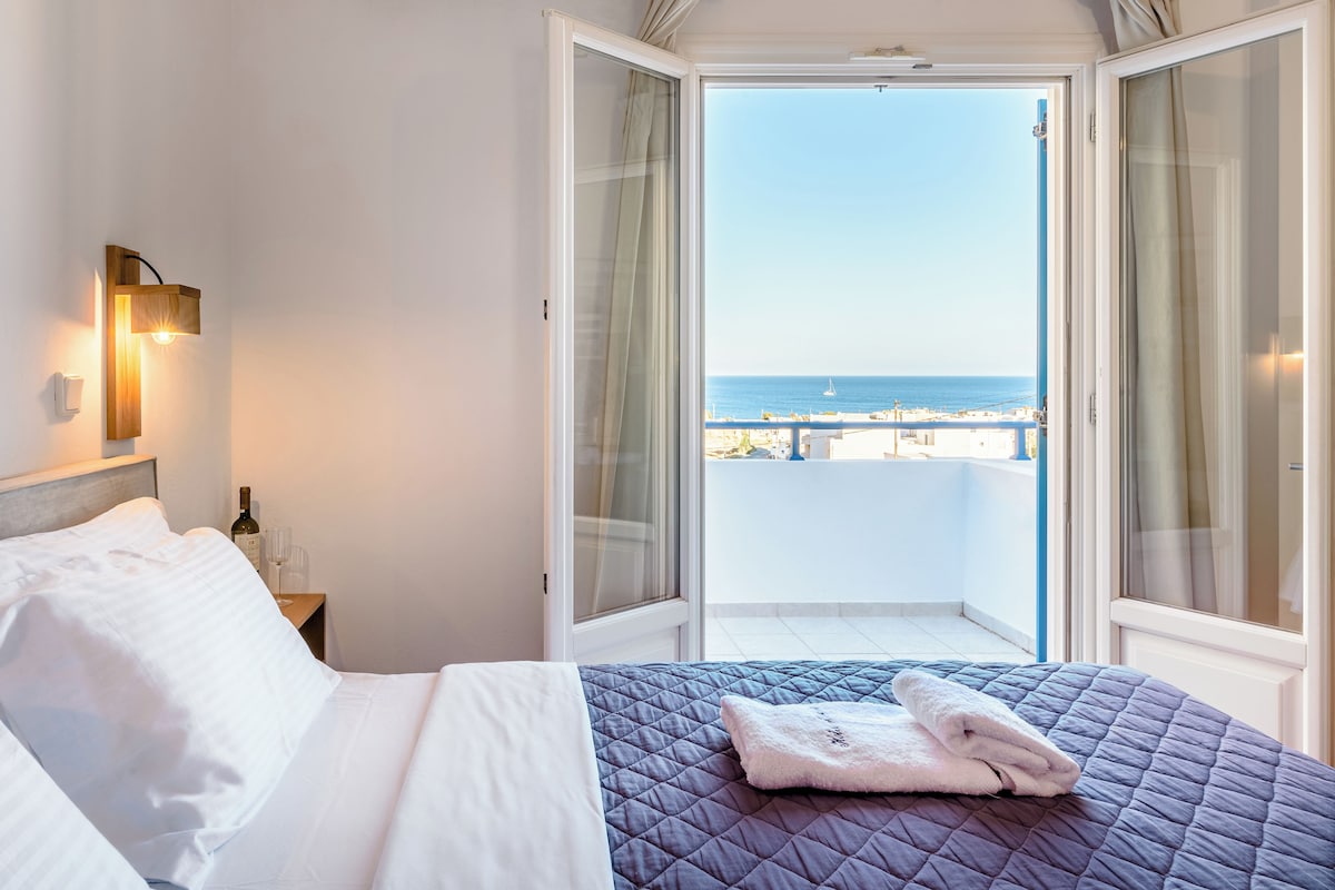 A hotel room with a bed, a nightstand, a bottle of wine, and a glass. There is a balcony with a view of the sea.