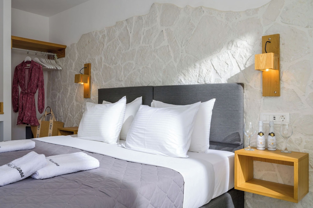 Cama bem arrumada com lençóis brancos, um edredom cinza e duas toalhas com texto. Há duas luminárias de parede, uma mesa de cabeceira de madeira com duas garrafas e copos, e uma parede de pedra.