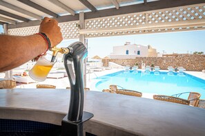 Eine Poolbar mit Zapfhahn, Korbstühle und Blick auf einen Swimmingpool sowie Gebäude im Hintergrund.