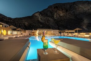 A poolside area with lounge chairs and a bar, overlooking a mountainous landscape. - Marianna Hotel (Santorini)