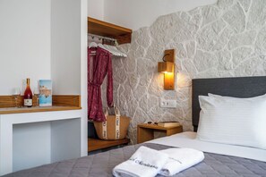A hotel room with a stone wall, a bed with white bedding, a wooden nightstand, a robe, a basket, and a bottle of wine. - Marianna Hotel (Santorini)
