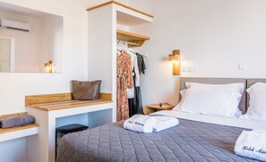 A hotel room with a bed, a wooden bench, a hanging clothes rack, a nightstand, and a wall-mounted light fixture. - Marianna Hotel (Santorini)