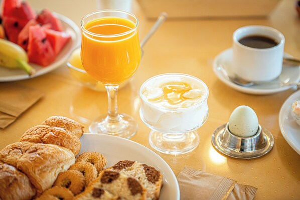 Une table dressée pour le petit-déjeuner avec des croissants, de la pastèque, du jus d’orange, du café et un bol de yaourt accompagné d’un œuf dur.