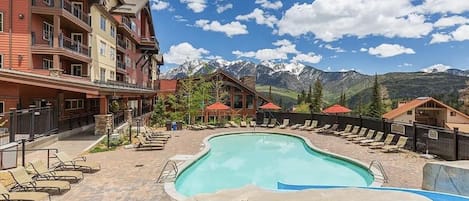 Uma área de piscina com espreguiçadeiras, vista para montanhas e um edifício com varandas.