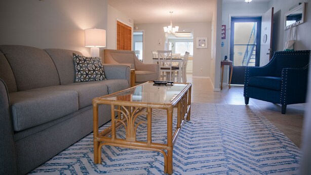 Een woonkamer met een bank, een salontafel en een blauwe fauteuil. Op de achtergrond is een eethoek met een kroonluchter.
