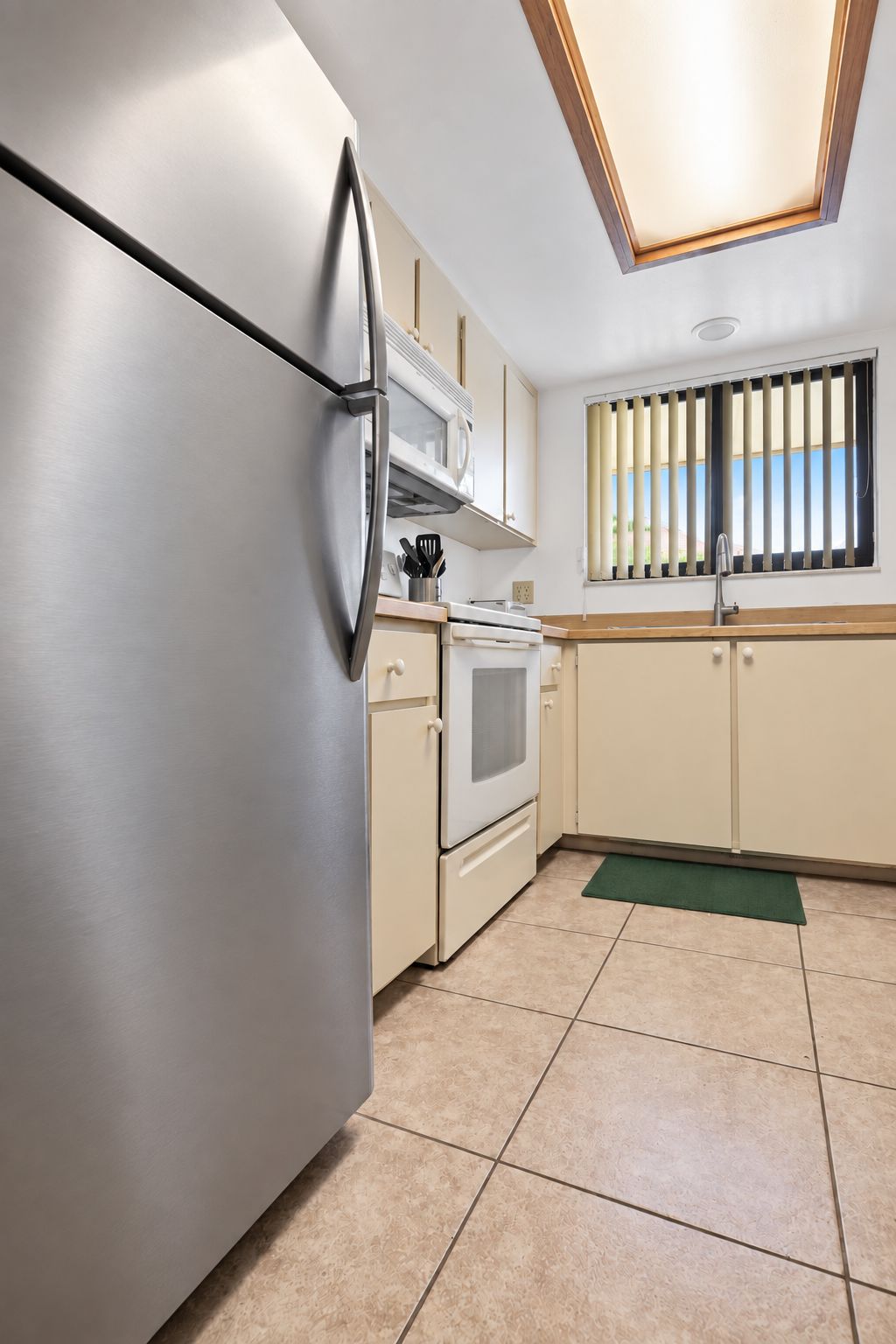Una cocina con un refrigerador de acero inoxidable, un microondas, un horno y una ventana con persianas verticales.