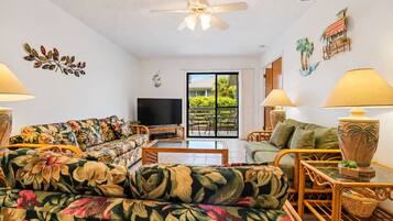 A living room with a sofa, a television, a table, and a lamp. There is a balcony with plants and a view of greenery.