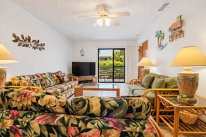 A living room with a sofa, a television, a table, and a lamp. There is a balcony with plants and a view of greenery. - 250 Sea Woods - 3BD - Your Beach Escape Awaits (New Smyrna Beach)