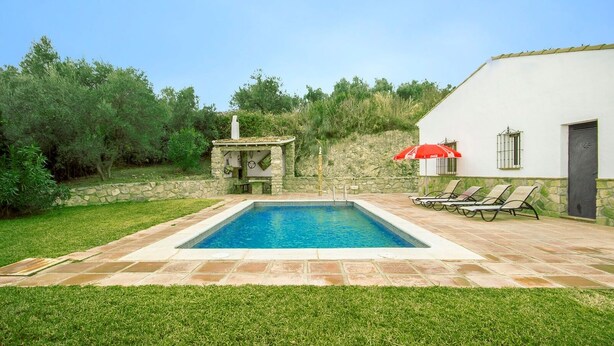Una piscina rectangular con borde de piedra, rodeada por un césped cuidado y un muro de piedra. Hay sillas de descanso y una sombrilla roja cerca de un edificio con puerta y ventanas.