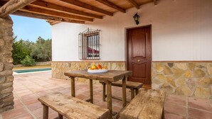 Un patio exterior con mesa y bancos de madera, pared de piedra, puerta de madera y ventana con barrotes de metal.