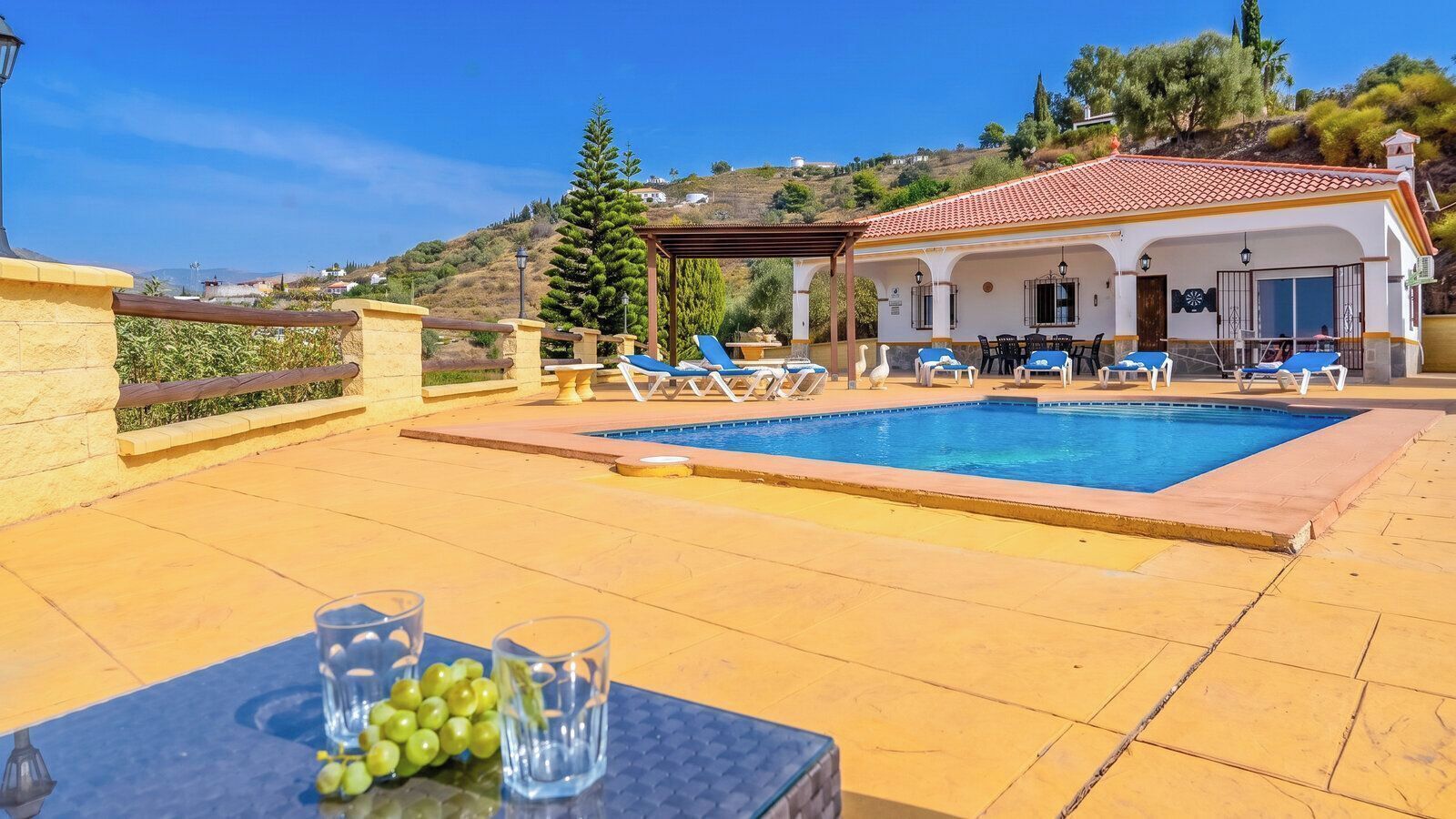 A poolside area with lounge chairs, a table with glasses and grapes, and a building with a tiled roof.