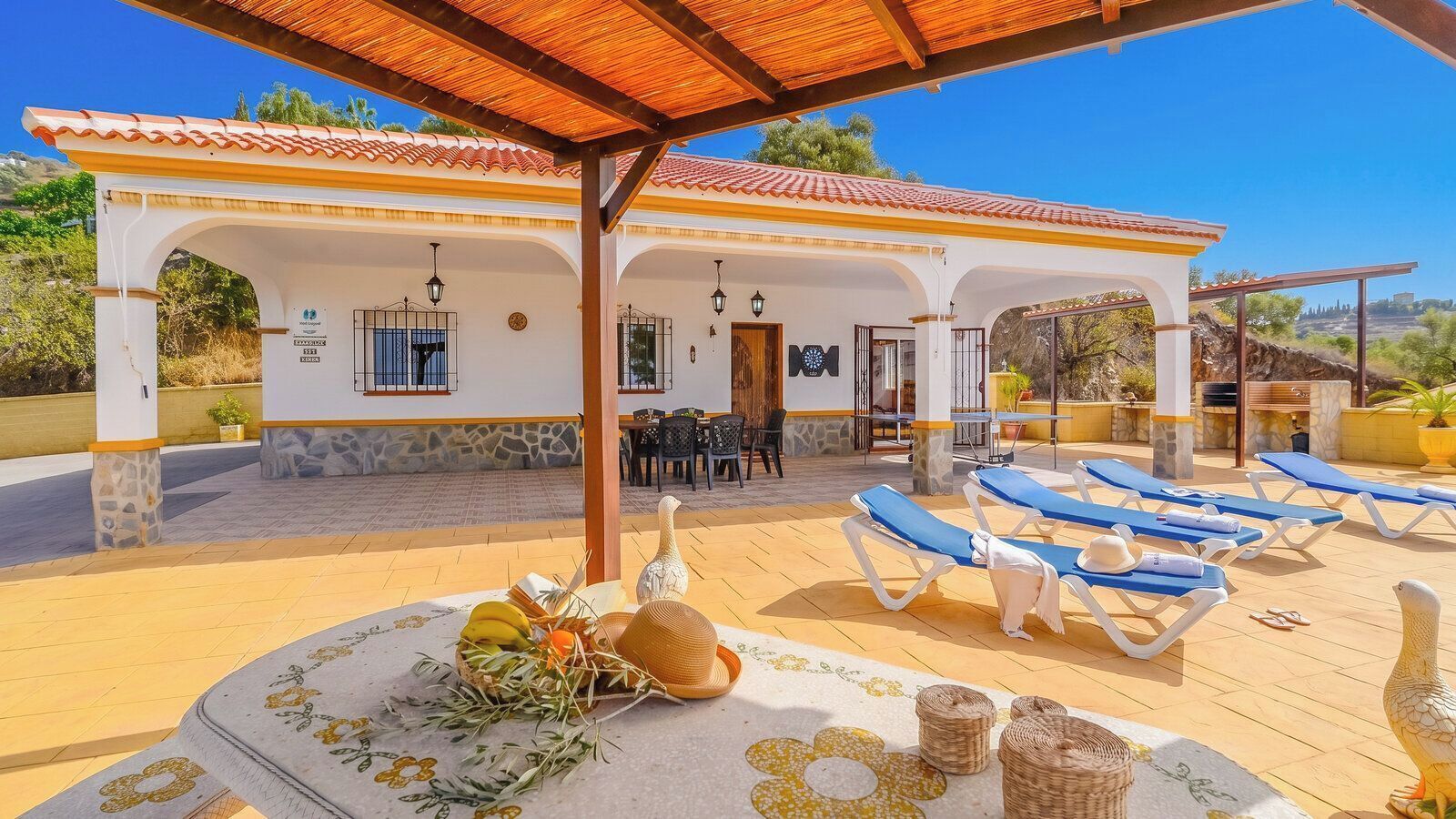 A covered outdoor area with a pool, lounge chairs, and a table with fruits and a hat.