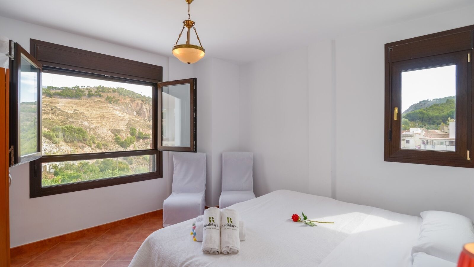A hotel room with a bed, two white chairs, a window with a view of a hilly landscape, and a chandelier.