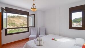 A hotel room with a bed, two white chairs, a window with a view of a hilly landscape, and a chandelier. - Casa Rural Parador del Silo Cónchar by Ruralidays (Granada)