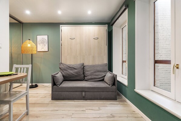 Een moderne woonkamer met een grijze bank, een eettafel met stoelen, een vloerlamp en een groot raam met uitzicht op een bakstenen muur.