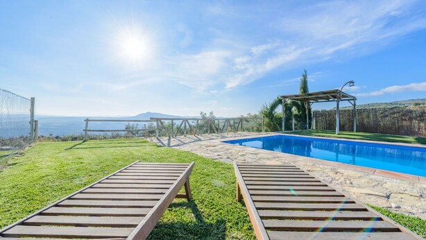 A poolside area with wooden loungers, a clear view of the sea, and a stone pathway leading to a wooden pergola.