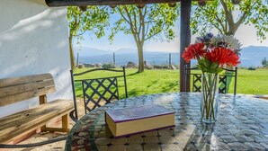 A table with a book and a vase of flowers, a bench, and a view of a grassy area and mountains.