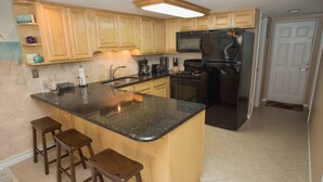A kitchen with wooden cabinets, a black refrigerator, a microwave, and a granite countertop. There are three wooden stools and a stainless steel sink. - Win San A-208 2BD - Sandy Toes & Salty Breezes (New Smyrna Beach)