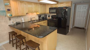 A kitchen with wooden cabinets, a black refrigerator, a microwave, and a granite countertop. There are three wooden stools and a stainless steel sink.